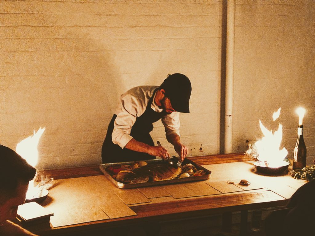 A chef preparing a large pan of food with flaming pans on either side of him