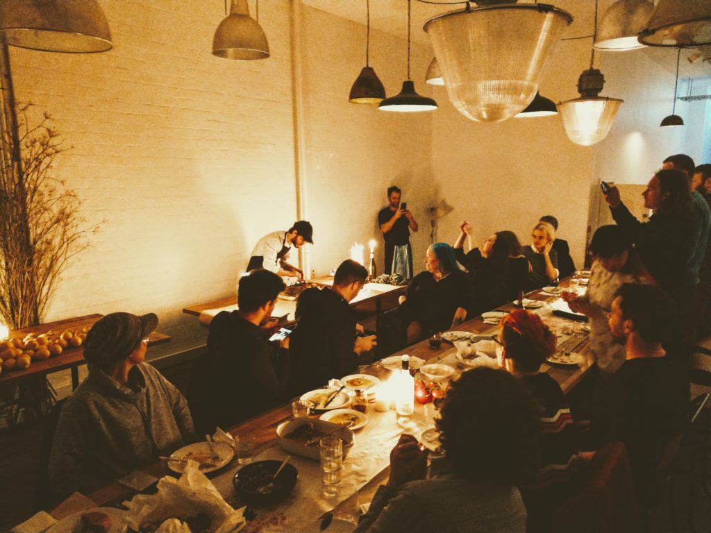 A group of people sat at long wooden table taking photos of a chef preparing food behind them