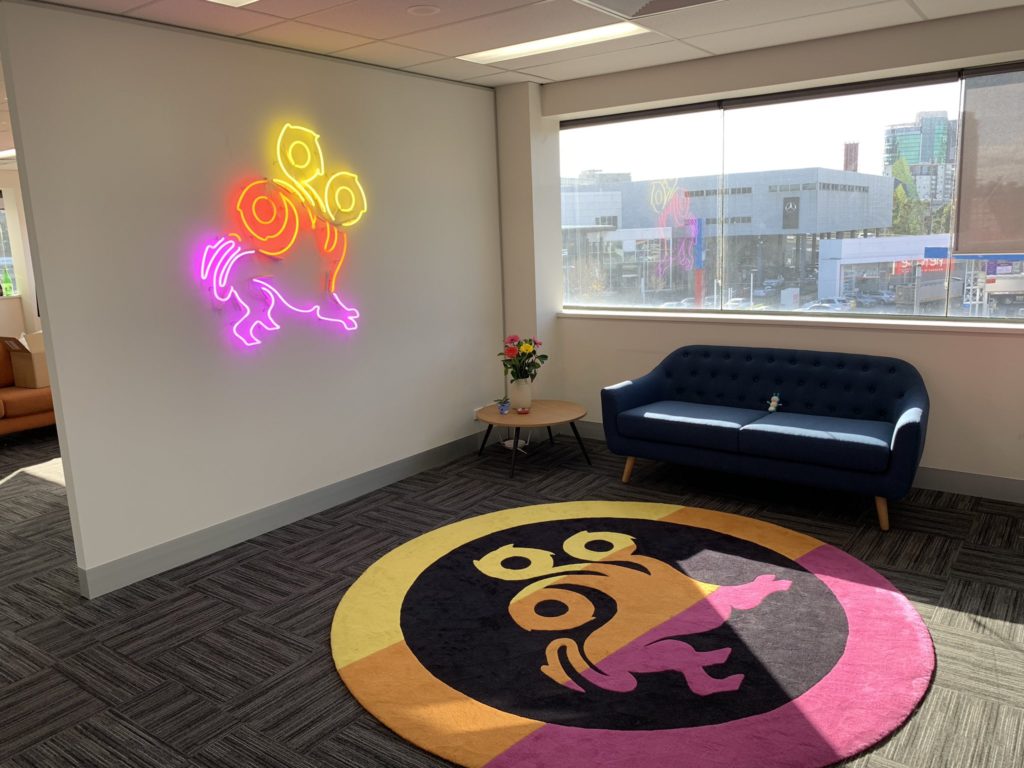 A wall with a neon sign version of the League of Geeks logo on it and a rug with the League of Geeks logo on it in front