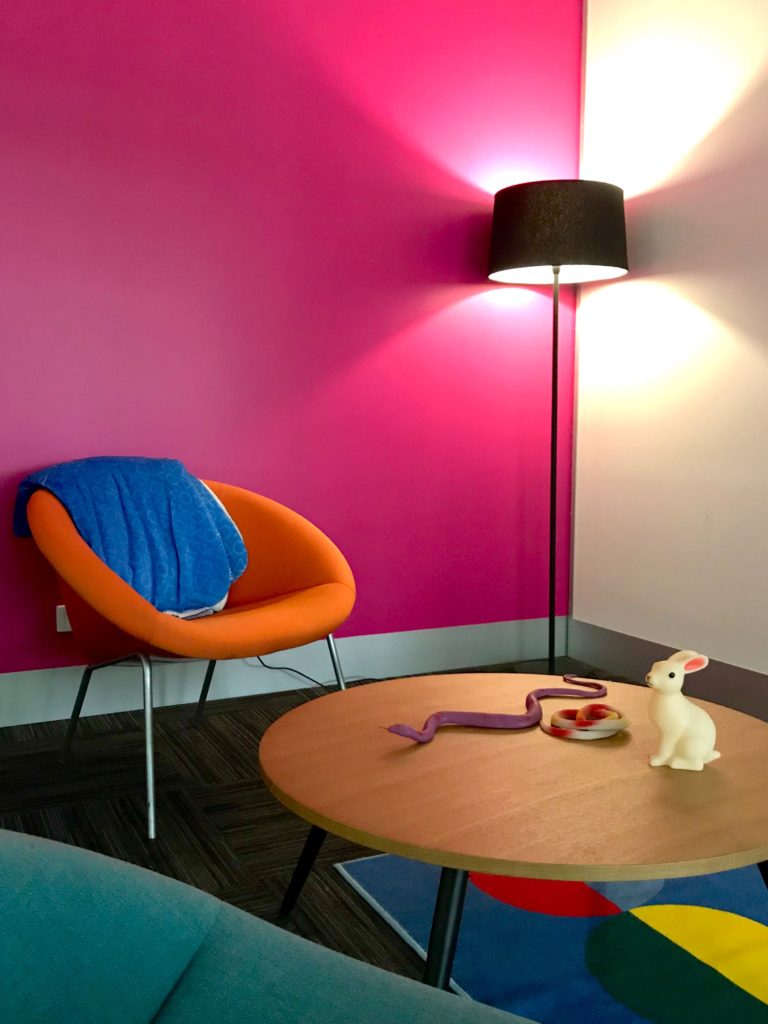 A breakout area of the office with a pink wall, orange chair, and a coffee table topped with a toy rabbit and snakes